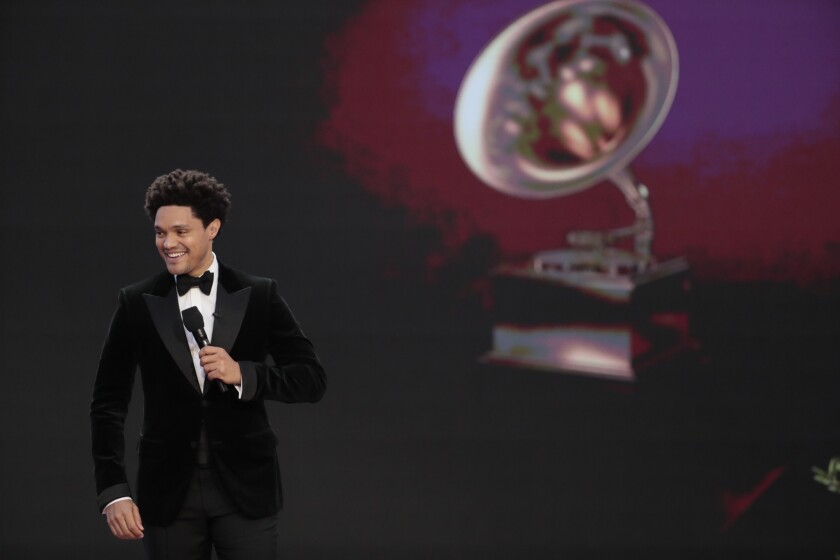 Trevor Noah holds a microphone while standing in front of an image of a Grammy