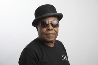 Tito Jackson poses for a portrait in a black t-shirt, dark aviator glasses and bowler hat