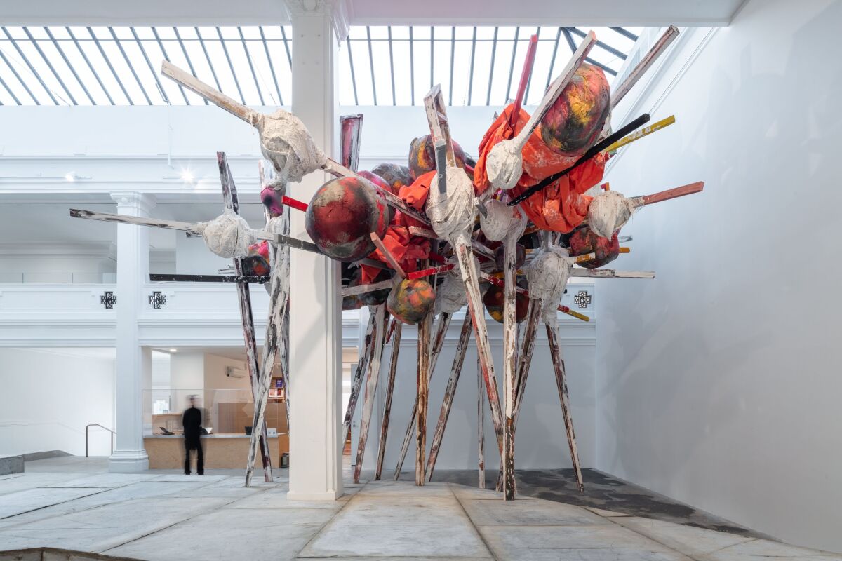A large sculpture with multiple wooden legs sits in a large white art space.