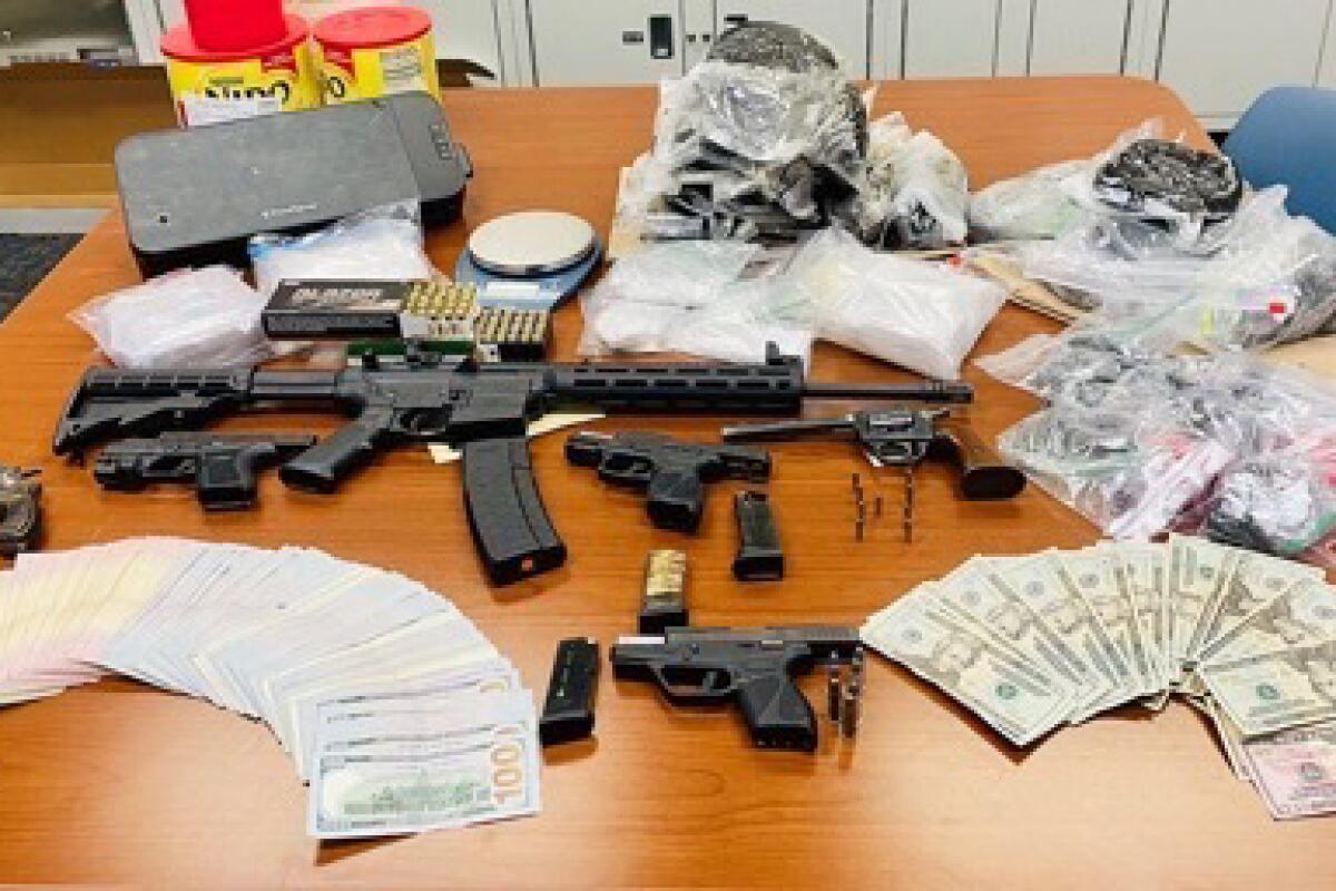 A table with plastic bags with heroin and fentanyl, spread US currency and an assault rifle.