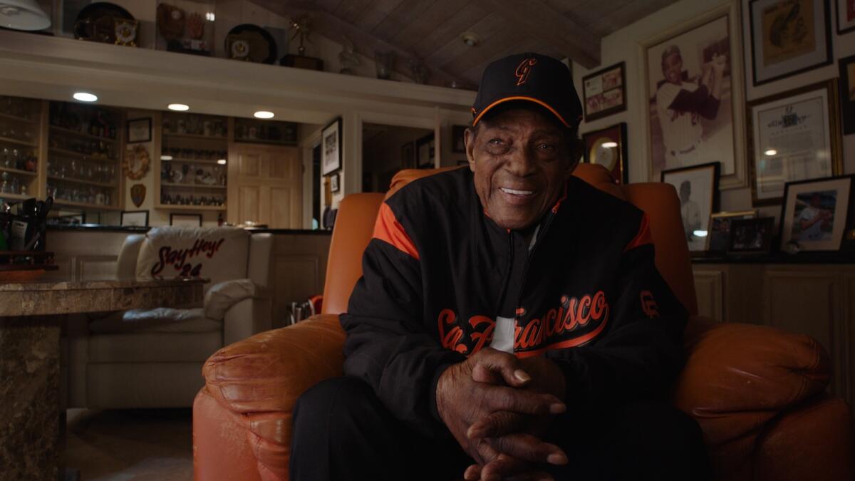 Willie Mays, wearing San Francisco Giants gear, sits in an overstuffed chair.