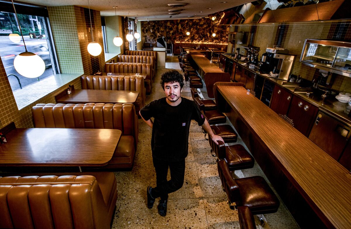 A man wearing all black stands in a traditional diner with a wood counter with stools on one side and booths on the other