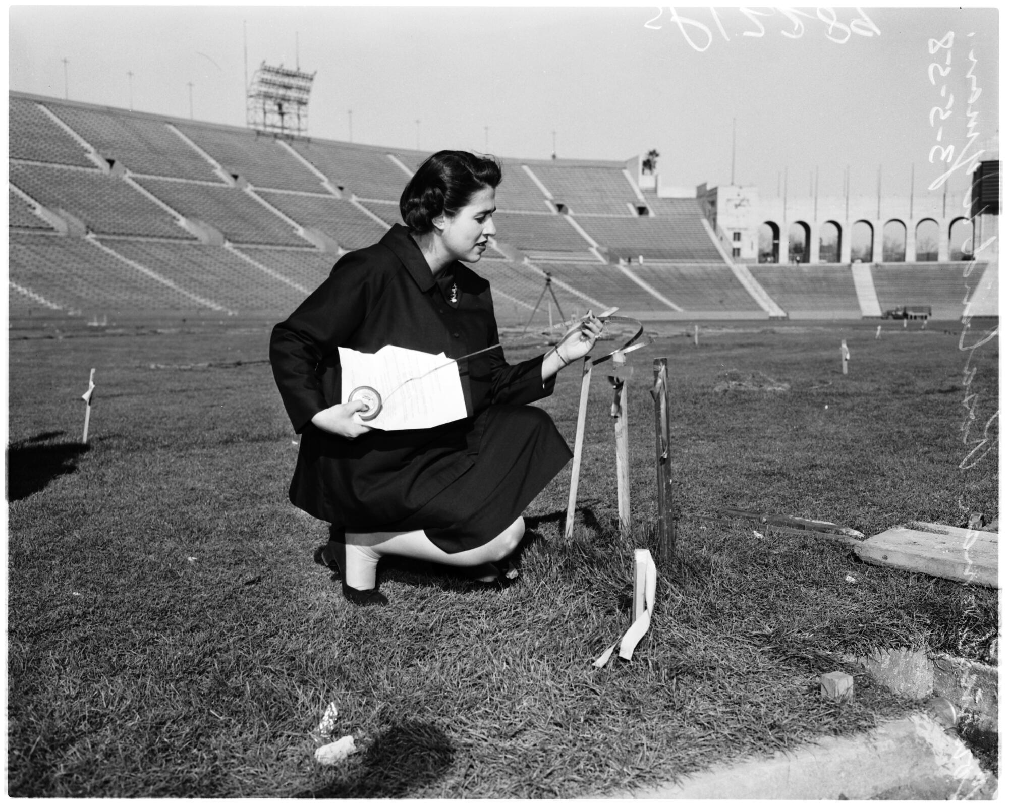A woman crouches near some sticks standing upright in the grass in a stadium.