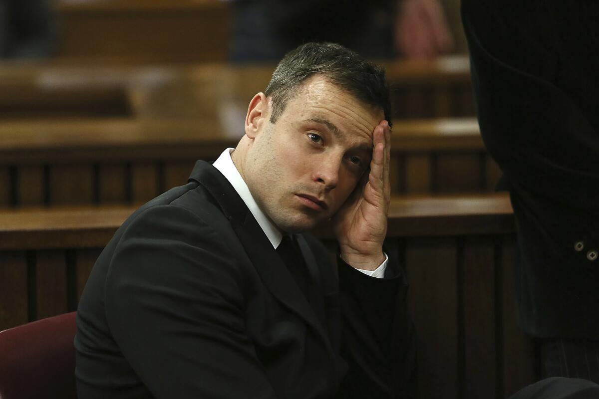 A man with short, dark hair, in a black suit, holds a hand to the side of his face as he sits in a courtroom 