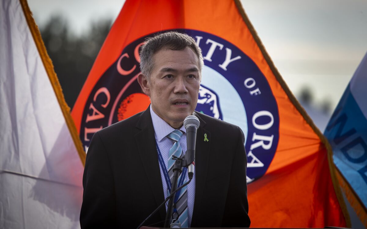 Dr. Clayton Chau speaks at a microphone in front of an Orange County flag