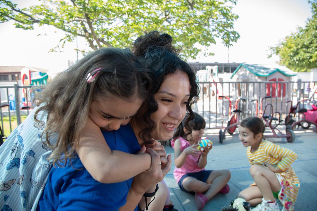 A teacher is hugged from behind by a preschool girl. 