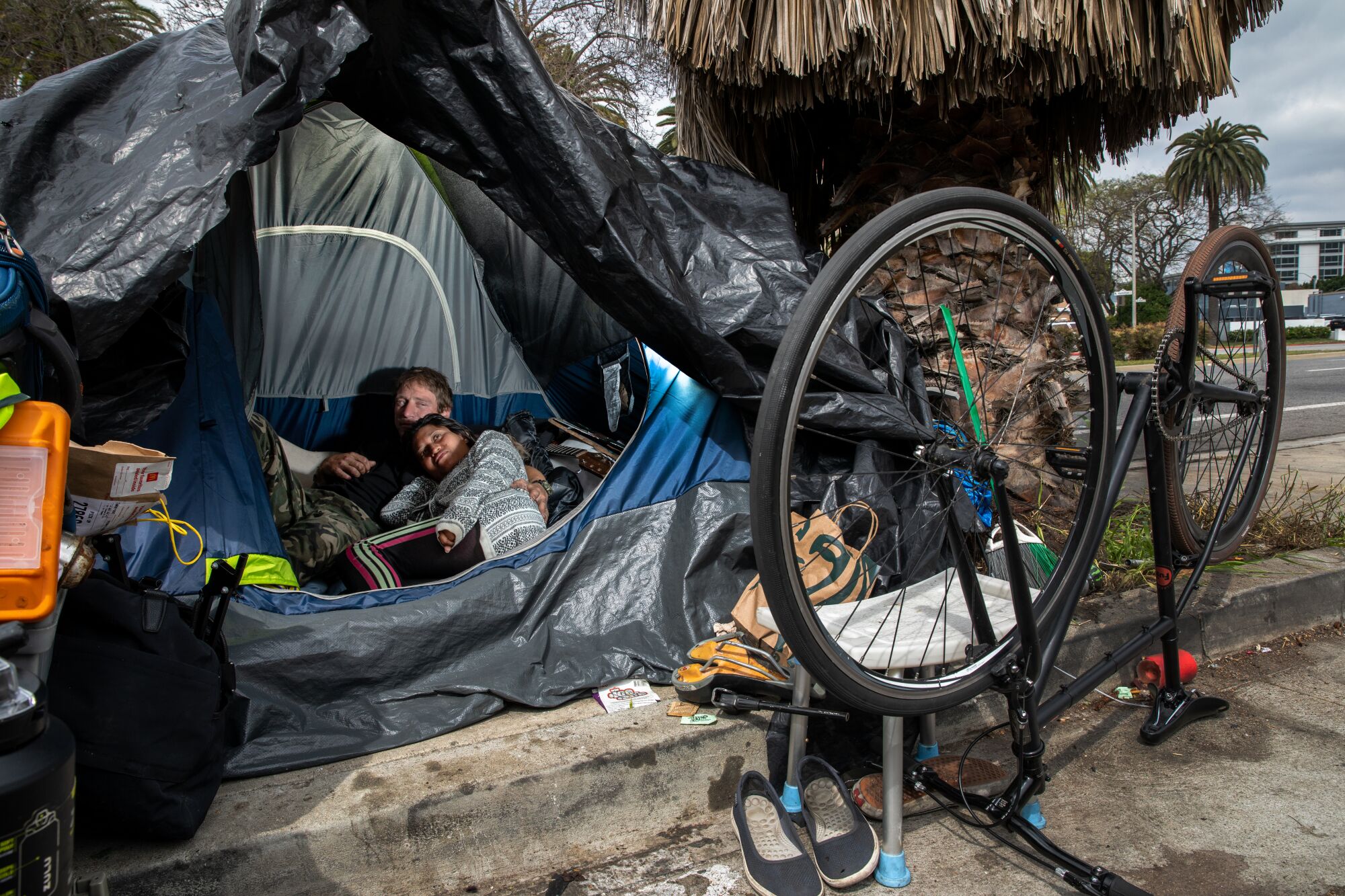 Two people lie in a tent beside a street with a bicycle and other belongings on the sidewalk