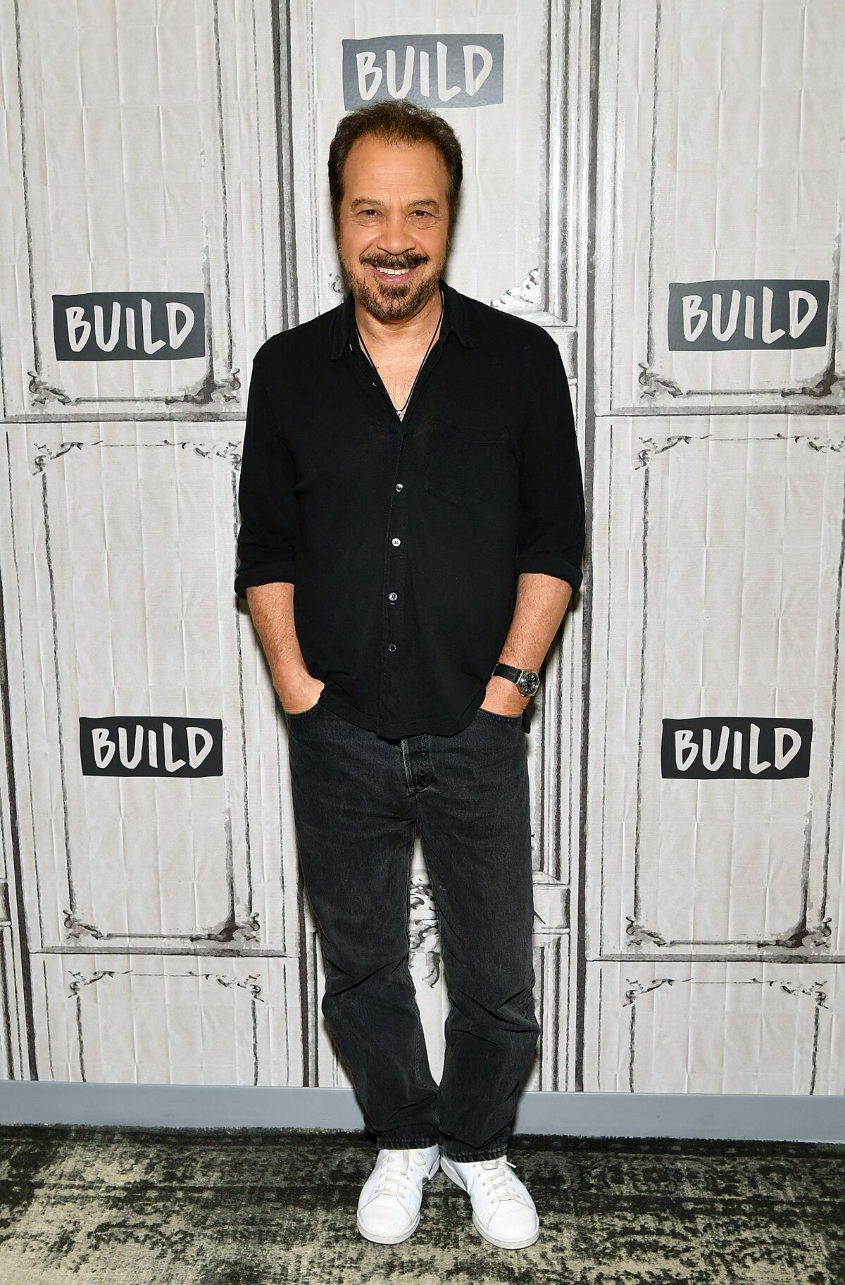 A man in a black shirt and pants smiles while standing with his hands in his pockets