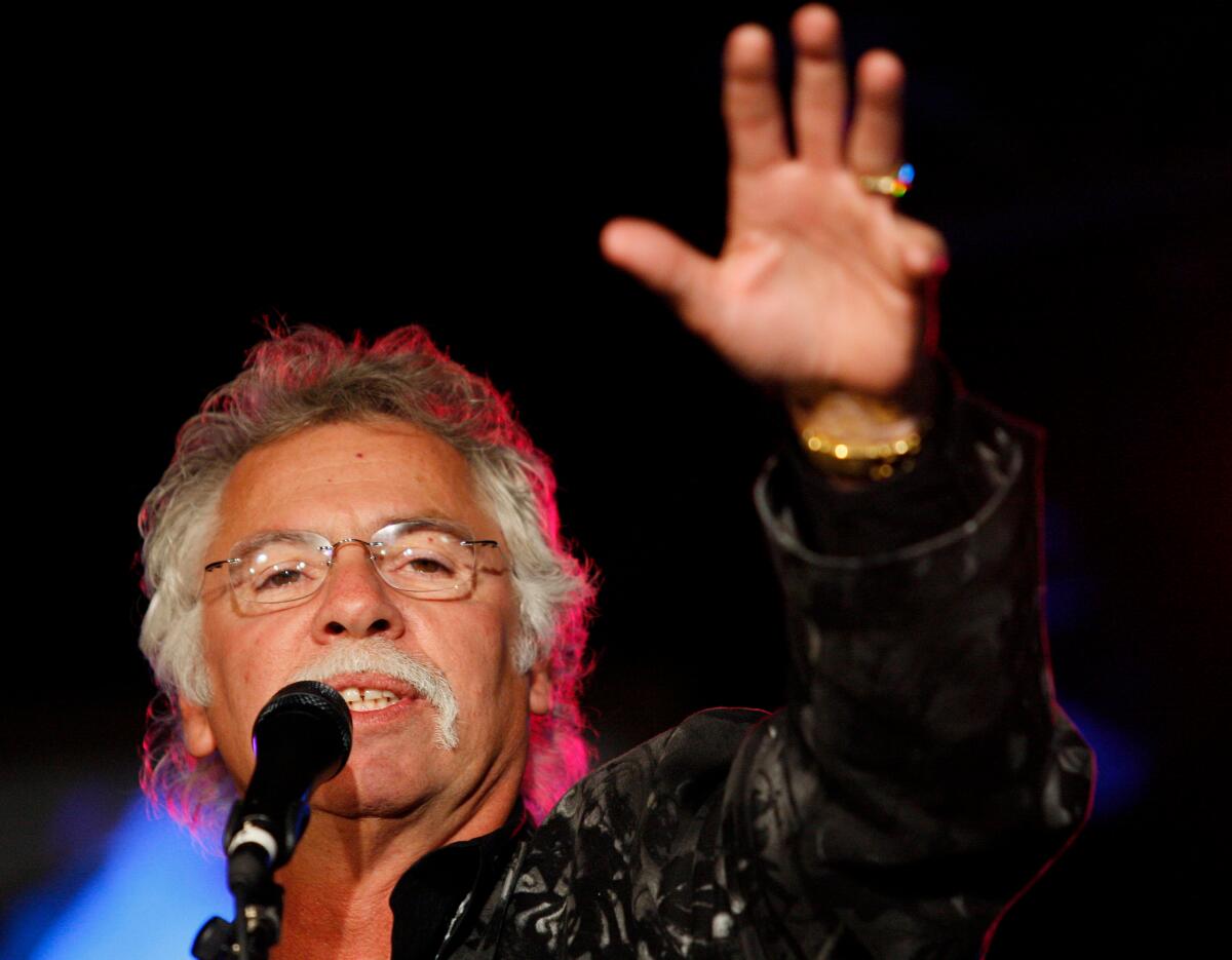 A man with graying hair, a mustache and wire-framed glasses singing into a microphone with his left arm raised