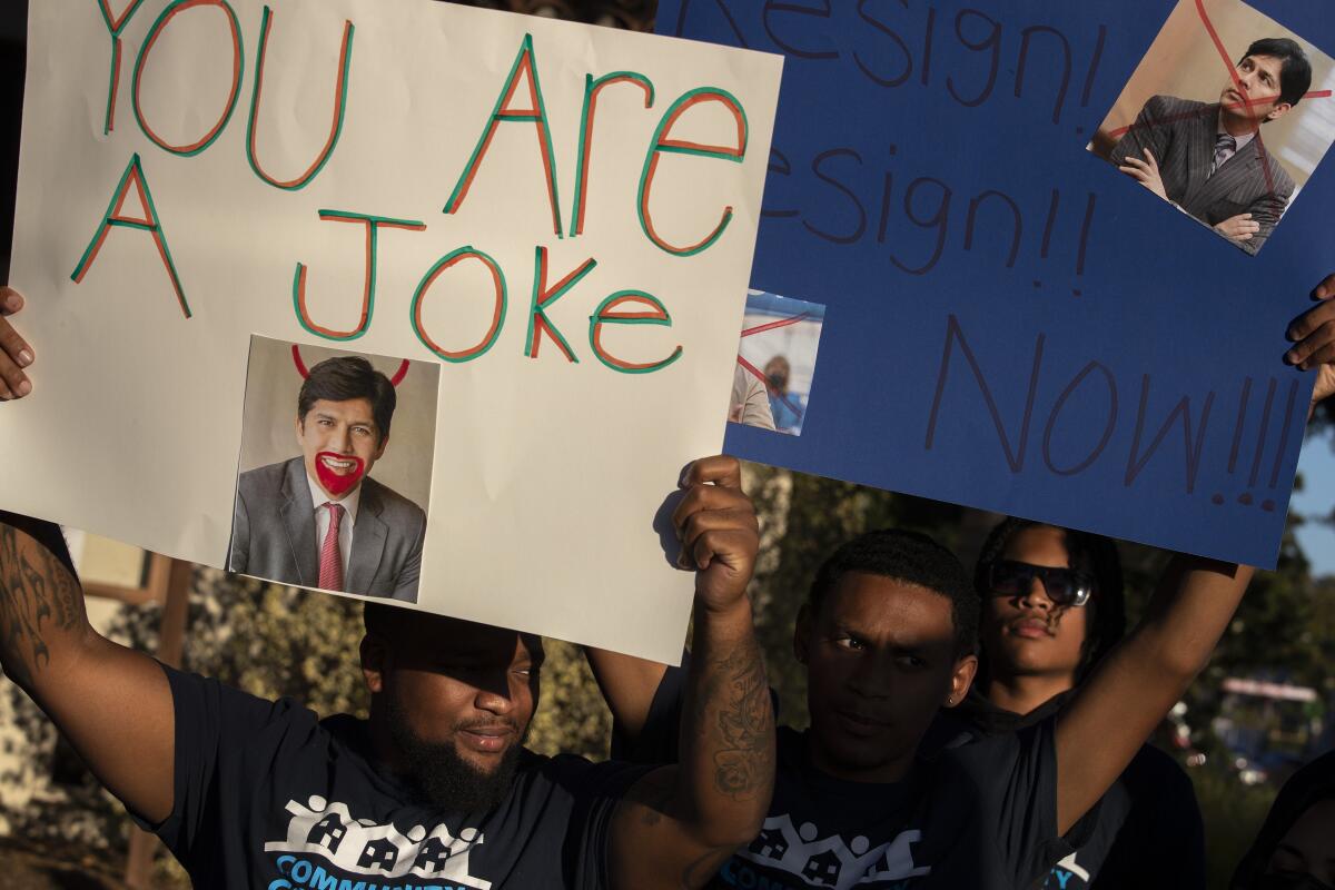 People hold up posters. One says "You are a joke," with a photo of a man with devil's horns drawn in. 