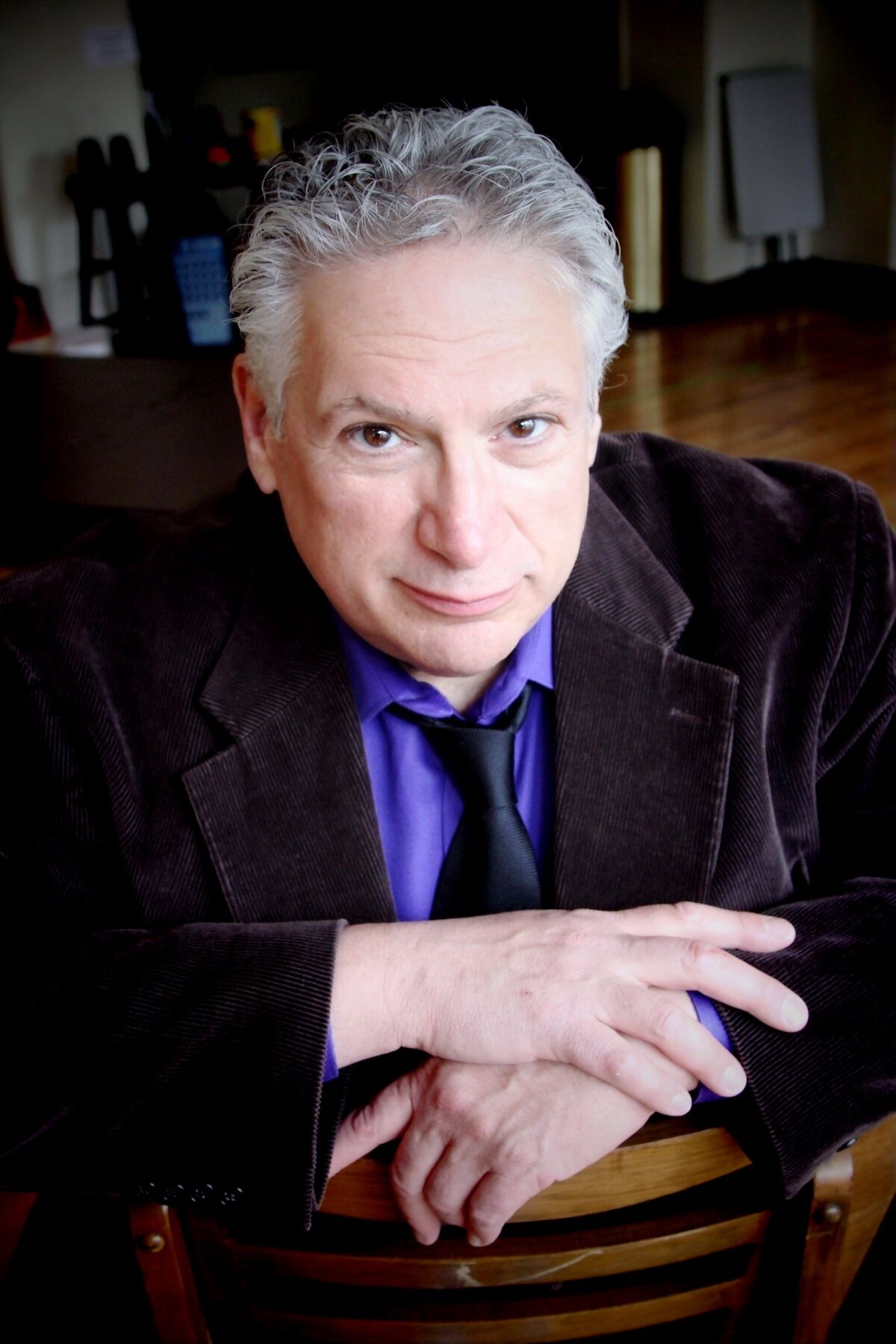 A gray-haired man in a plum velvet jacket sits with his hands on the back of a chair.