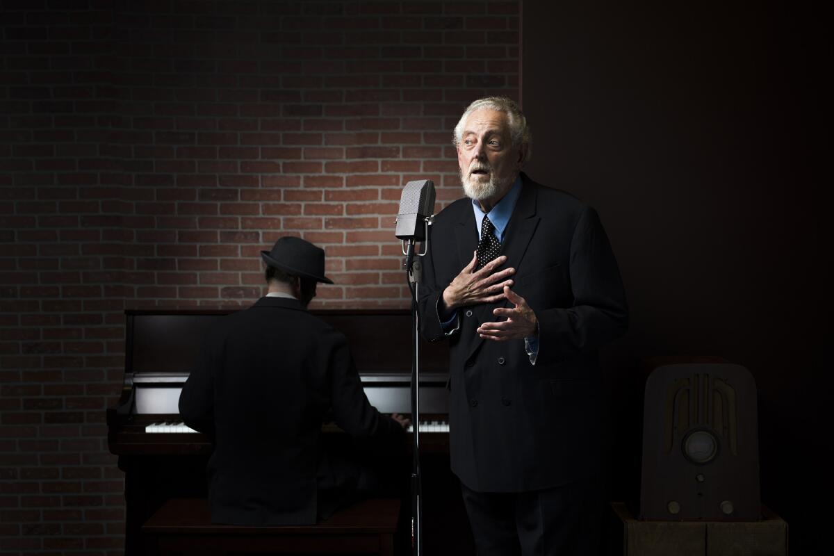 An elderly man in a nice suit speaks into a microphone on stage while a piano player plays in the background