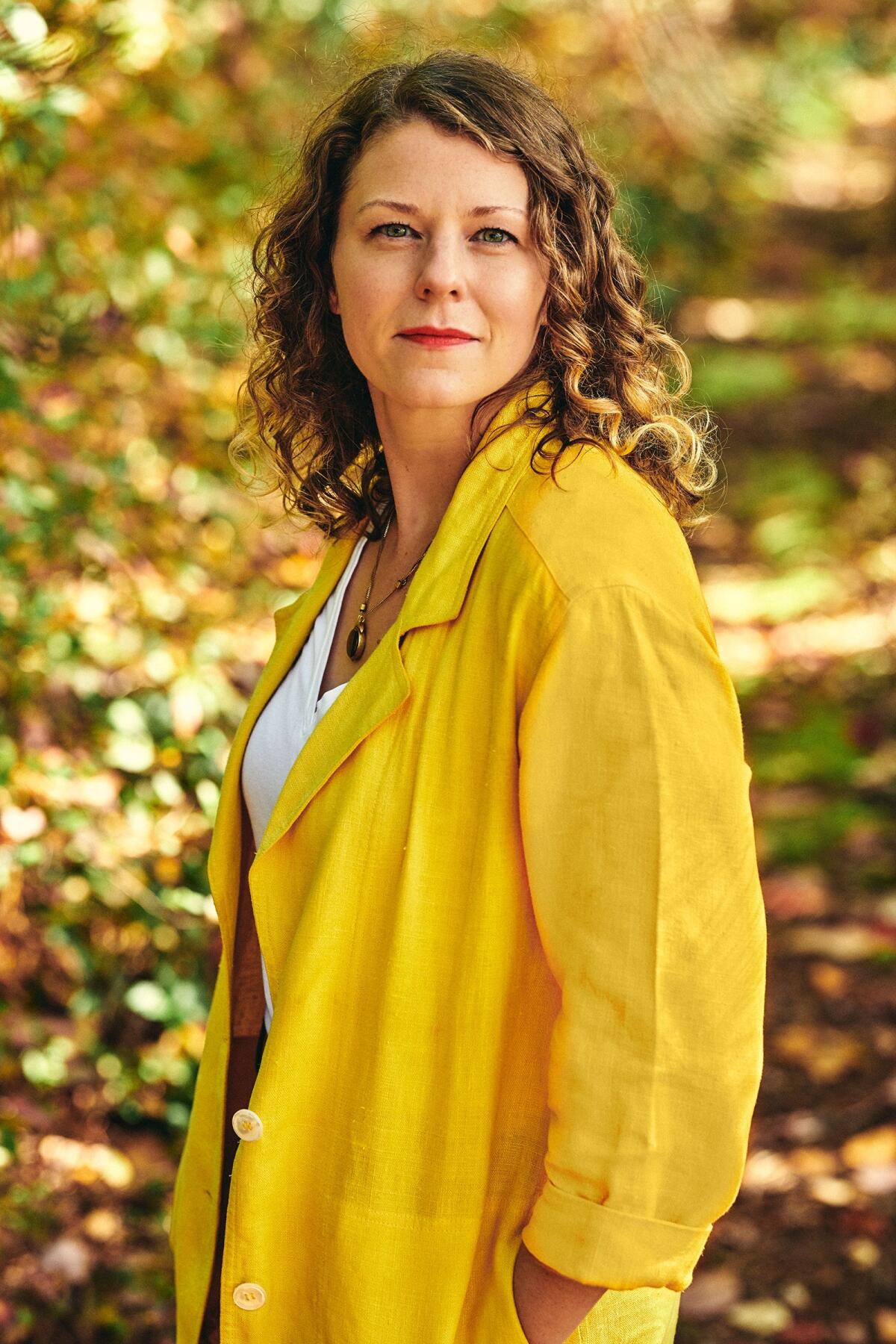 A woman in a yellow jacket stands in front of greenery.
