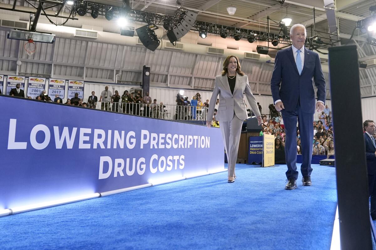 Vice President Kamala Harris and President Biden walk by a blue banner that reads "Lowering prescription drug costs."