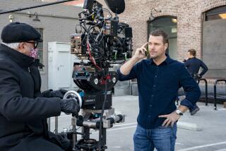 A man wearing a jacket, face mask and a hat filming another man in a collared shirt holding a phone to his ear on a film set