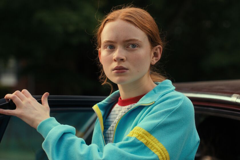 A girl with red hair wearing a blue jacket and holding a car door open
