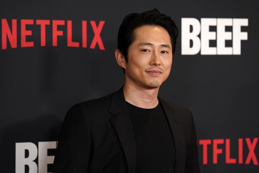 Steven Yeun in a black shirt and black blazer posing for pictures against a black backdrop with red and white text