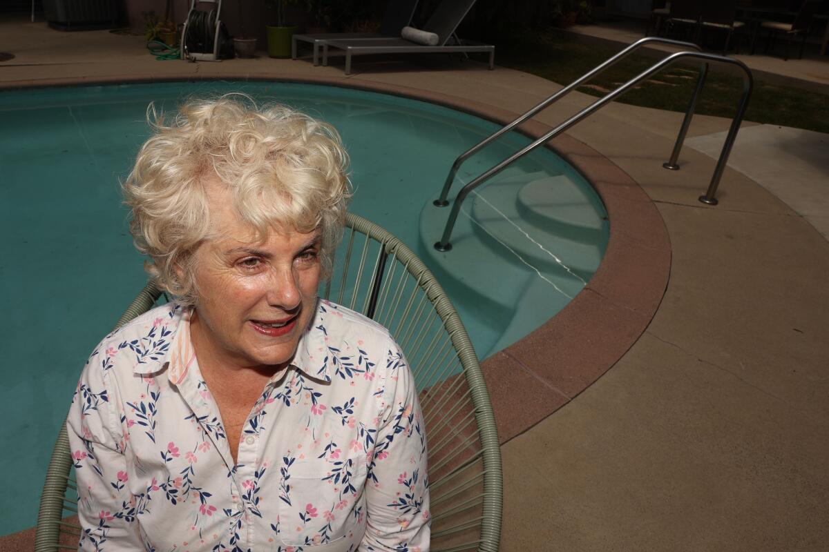 A woman in a flowered button-up shirt and curly blond hair sits near an oval swimming pool.