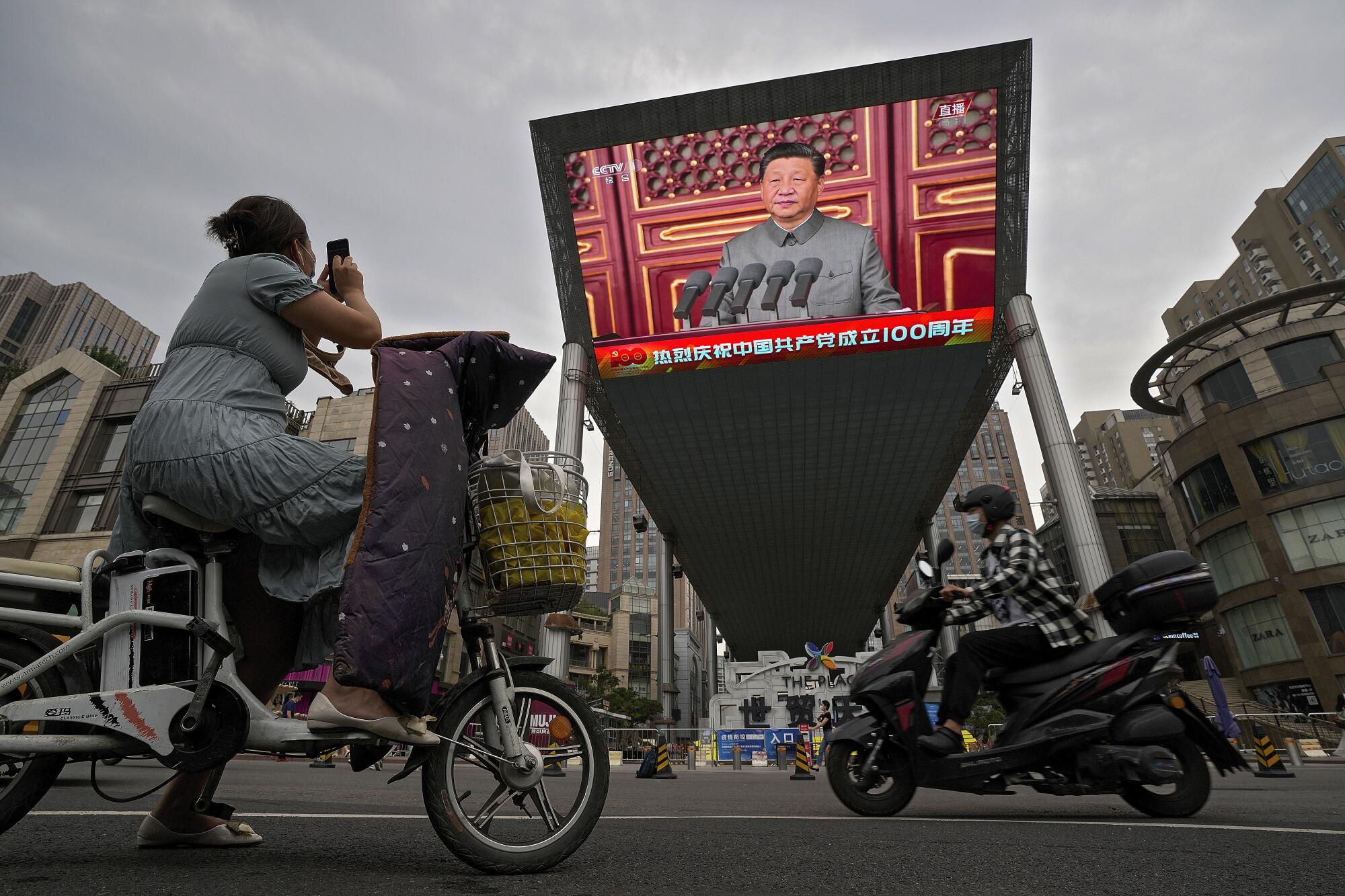 A woman on her electric-powered scooter films a large video screen showing Chinese President Xi Jinping.