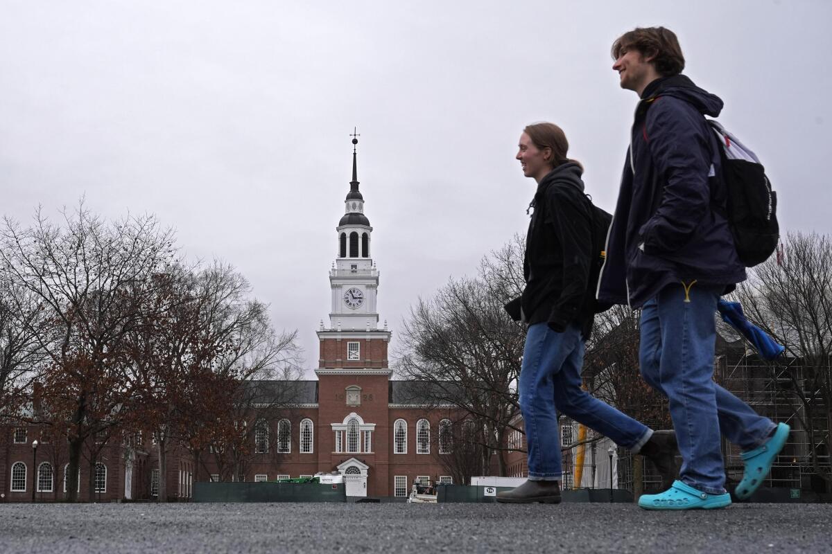Two young people with backpacks walking under a gray sky, bare trees and a brick building with a clocktower in the background
