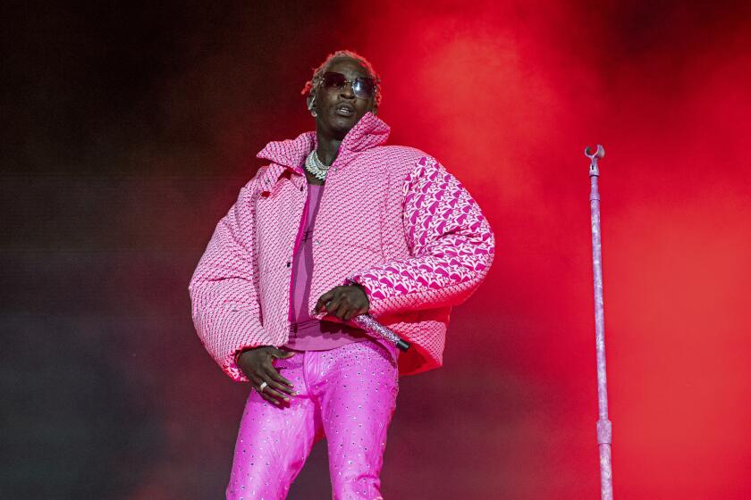 Young Thug wearing an oversized pink puffer jacket and tight hot pink pants on a stage. He stands by a mic stand