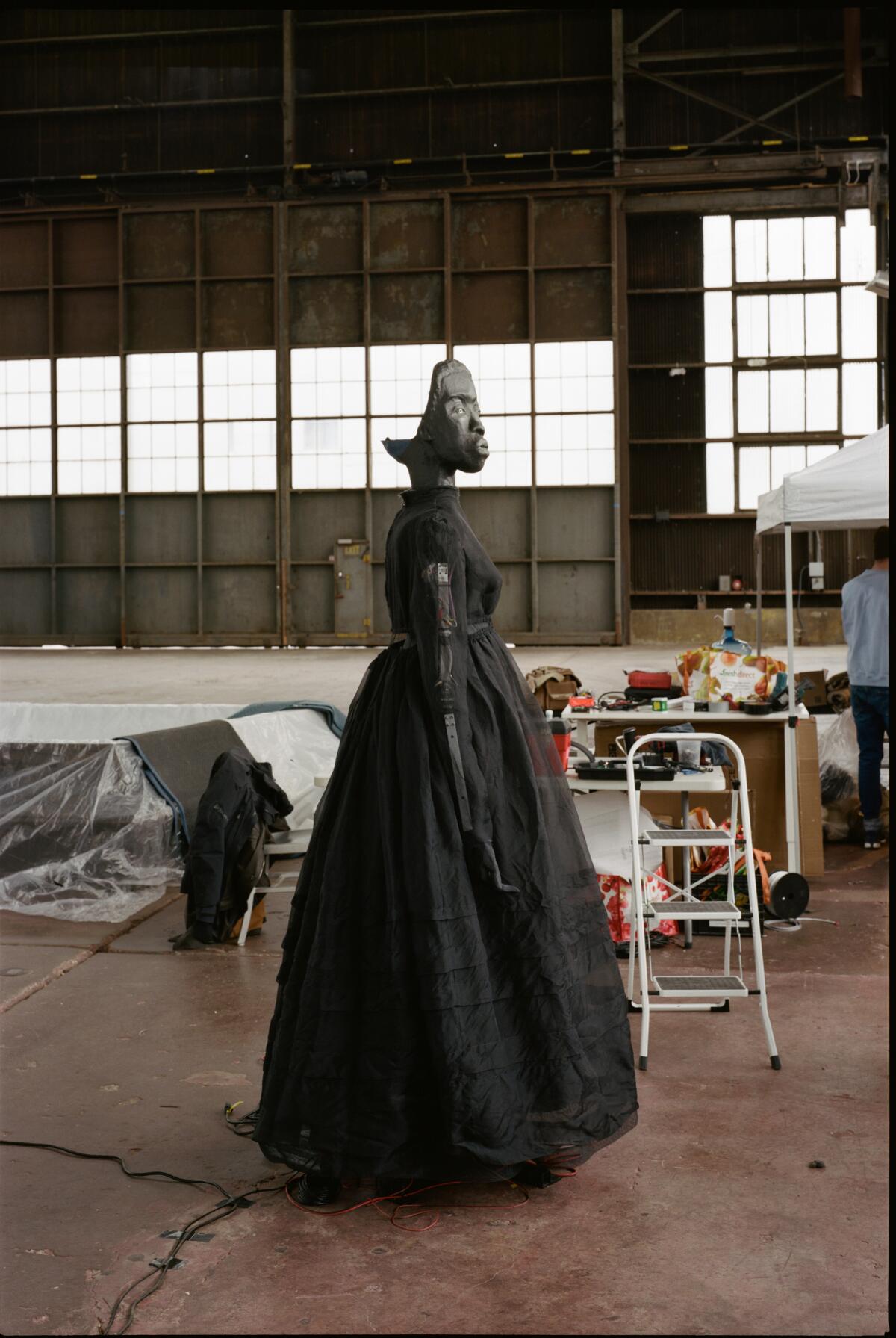 A Black mask-like face connects to a life-size female figure in a black dress, part of an in-progress installation.    