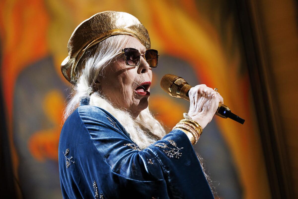A woman with gray hair, weiring a gold beret and dark glasses, sings into a handheld microphone onstage