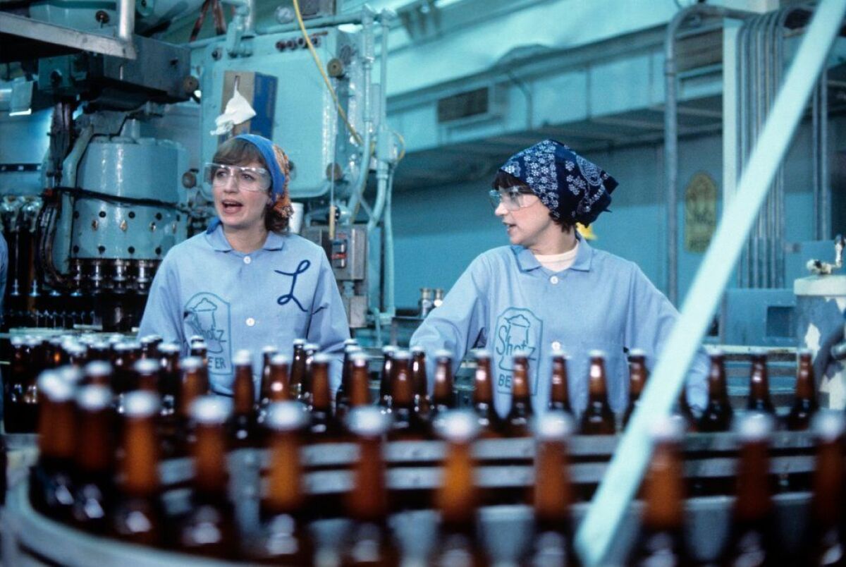Two women in 1950s clothing stand next to a beer bottle production line.