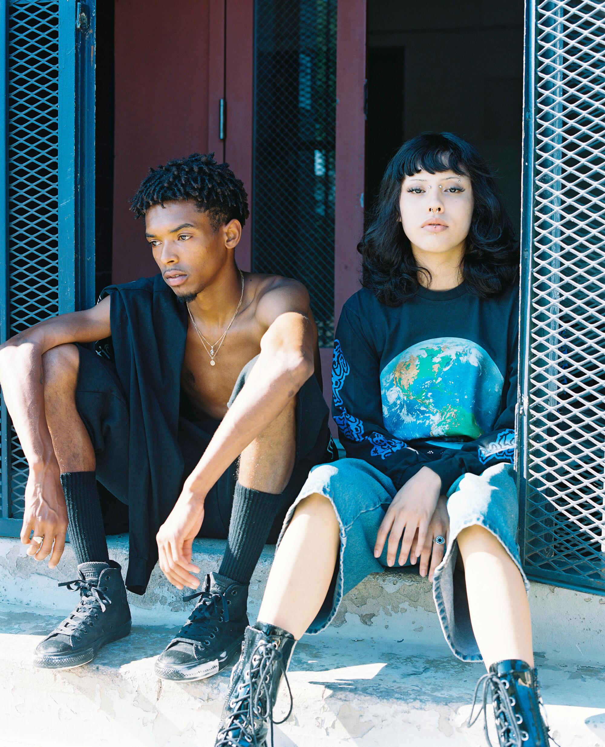 Two people sit casually on the sidewalk, wearing Total Luxury Spa outfits.