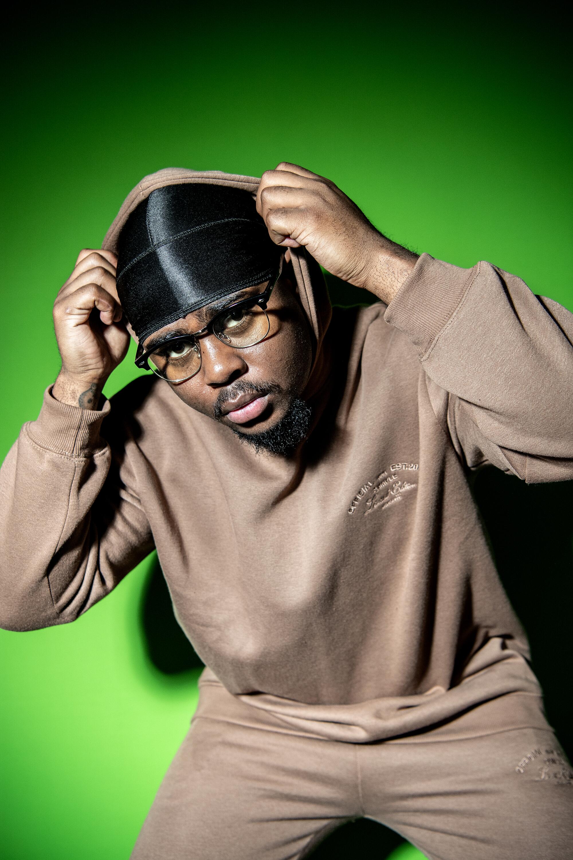 A man in a brown sweatsuit and a durag in front of a green screen 