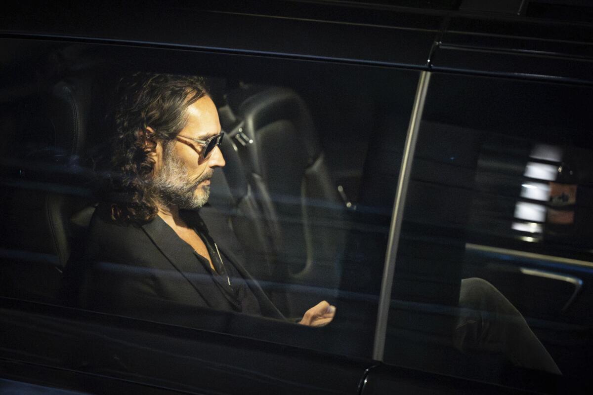 Russell Brand wears all black and sunglasses as he is seen from the side riding in the backseat of a black car