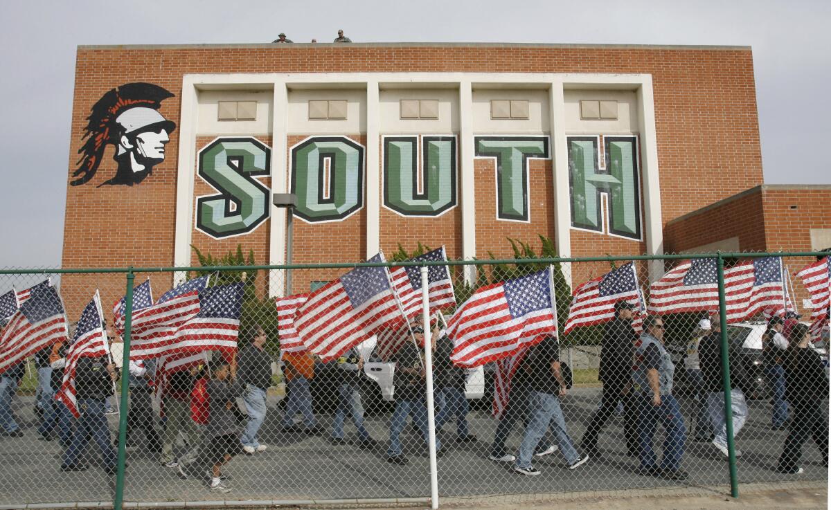 Several people carrying U.S. flags walk in front of a school building with the word "South"