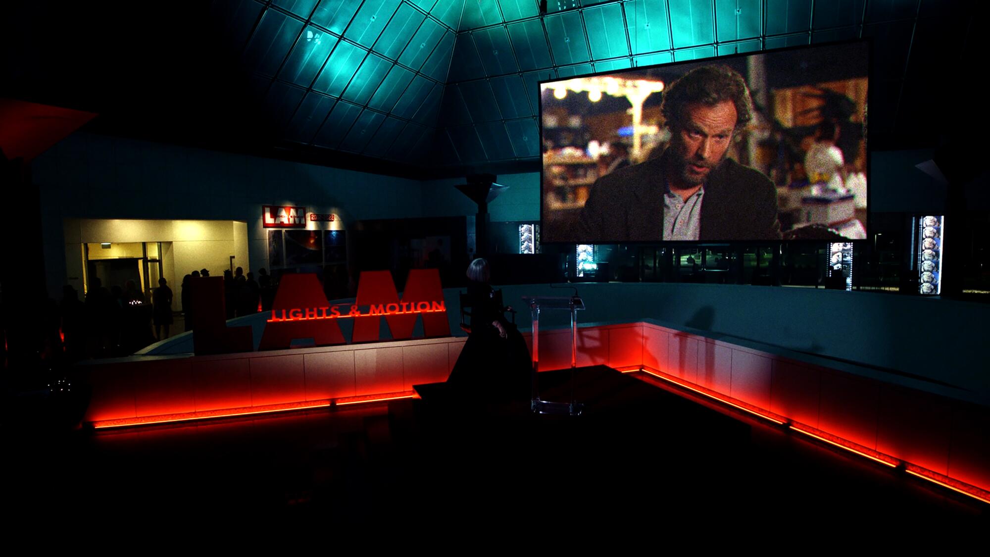 A large darkened gallery-type space features a huge TV screen displaying a man talking in a scene from "Poker Face."