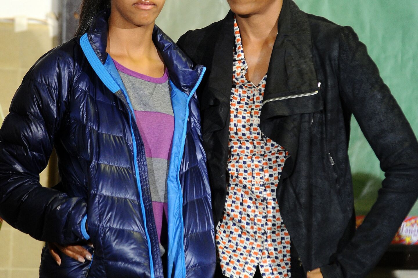 First Lady Michelle Obama, in a casual print top, textured leather jacket and dark jeans, and daughter Malia listen as President Obama speaks to volunteers at the Browne Education Center in Washington in January of 2012.