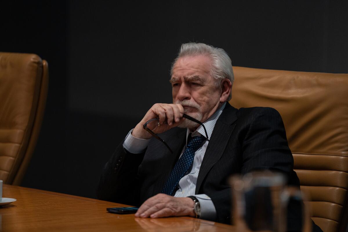 Brian Cox holds a pair of glasses in his hand while sitting at a table in a boardroom. He wears a black suit and tie