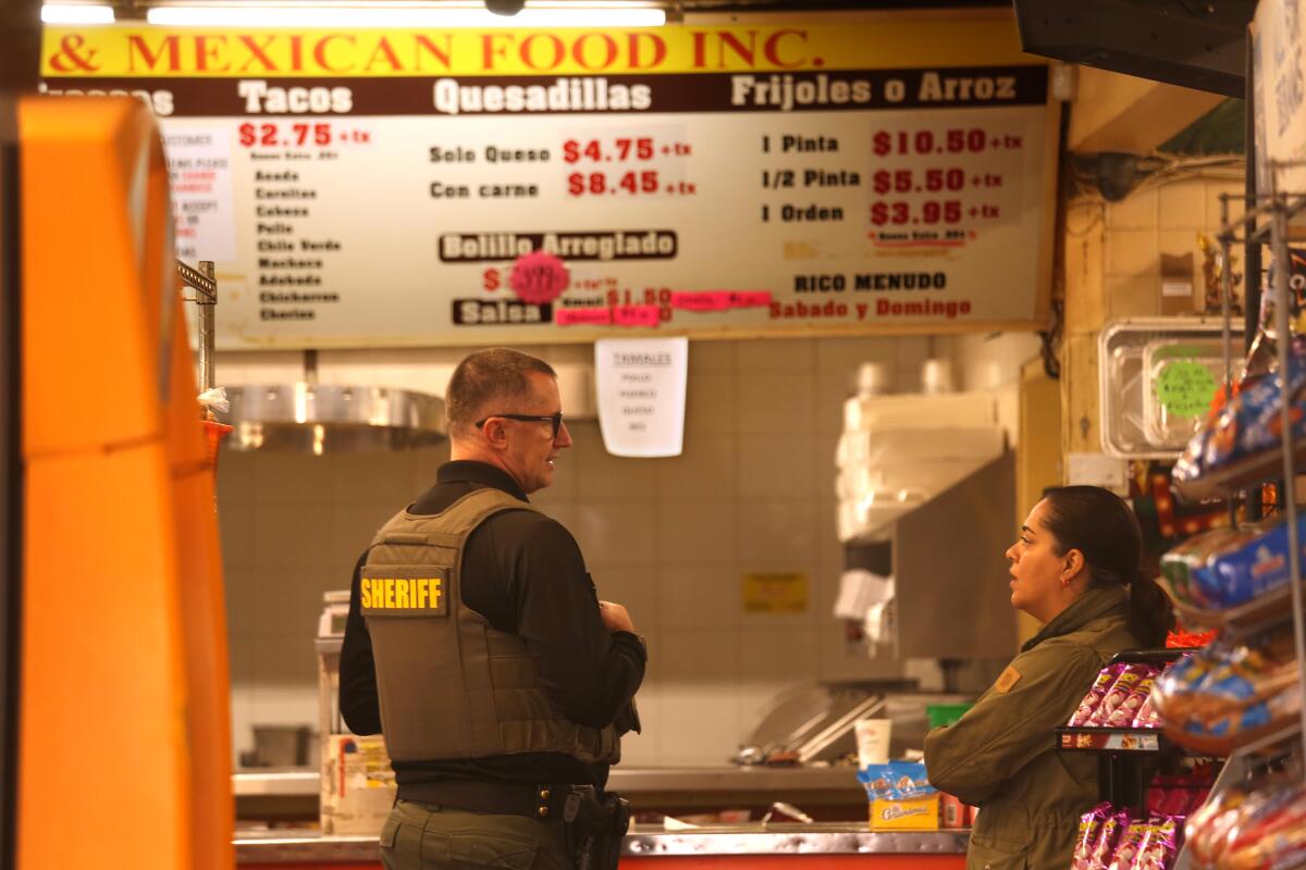 A man, left, wearing glasses and an olive vest with the word Sheriff speaks to a woman inside a store 