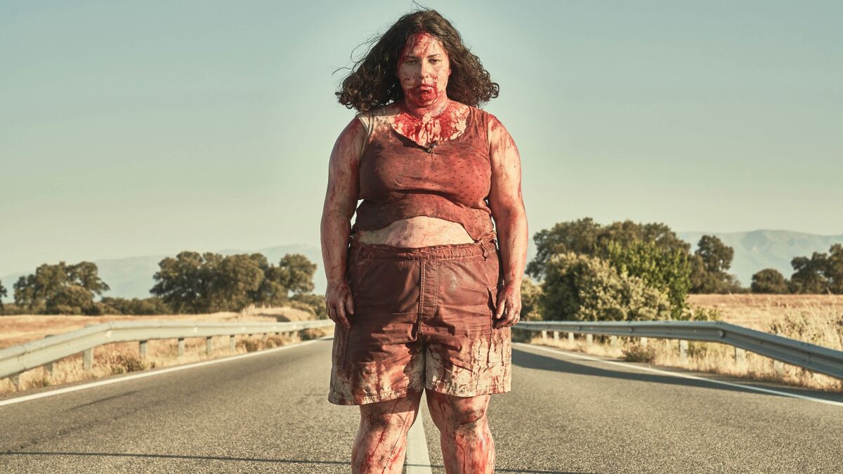 A woman covered in blood stands in the middle of a road 