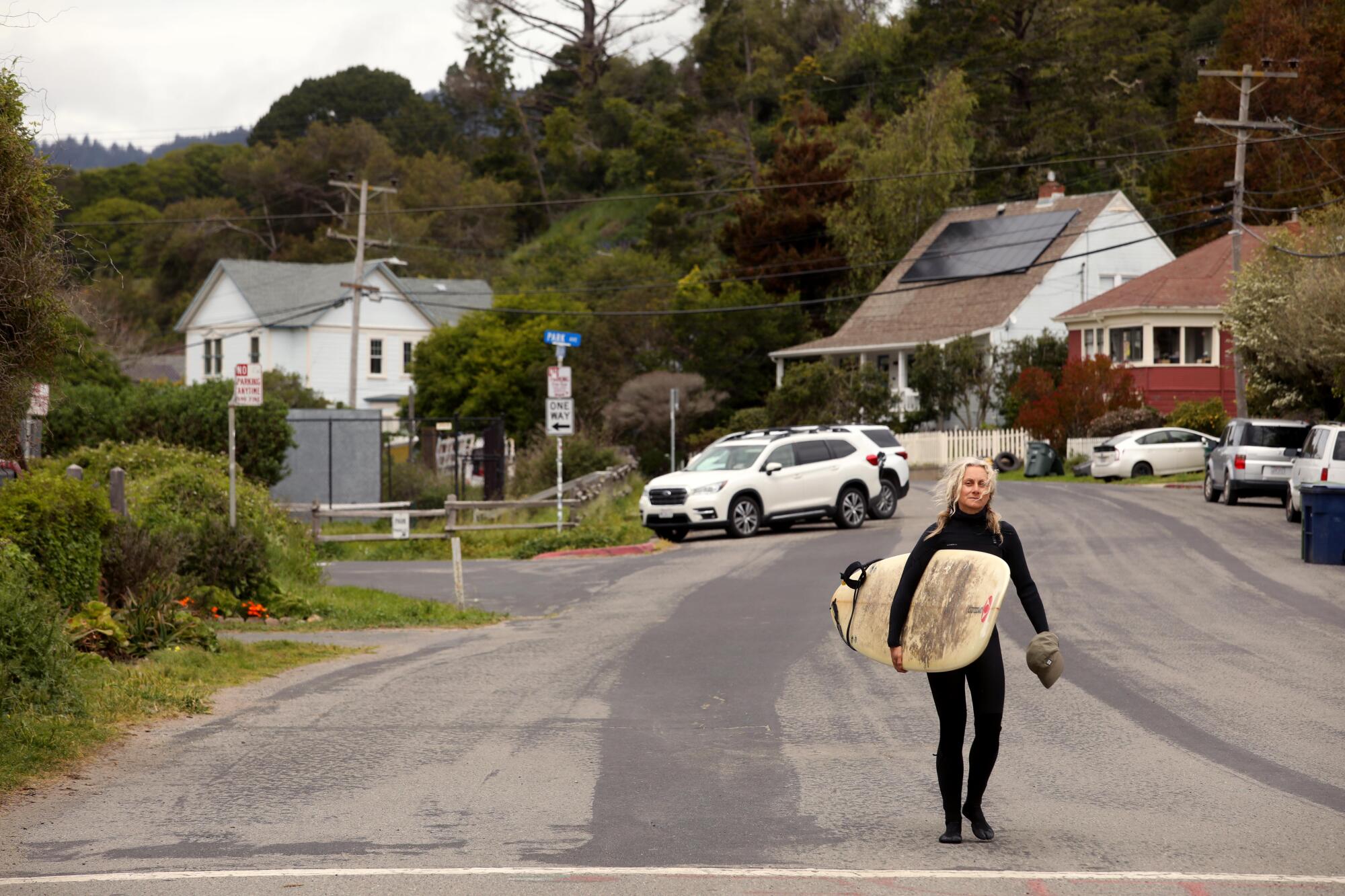 Uma mulher com roupa de neoprene preta carrega sua prancha de surf por uma rua vazia.