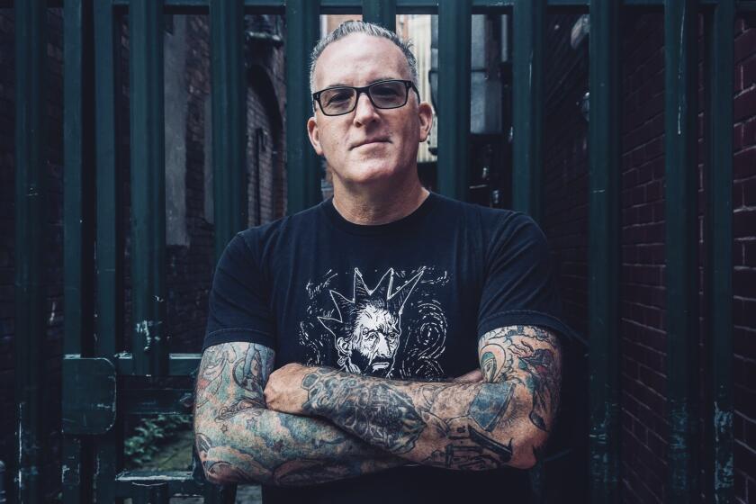 Man in dark glasses with a band shirt and heavy tattoos on his arms, standing in front of a gated alley.