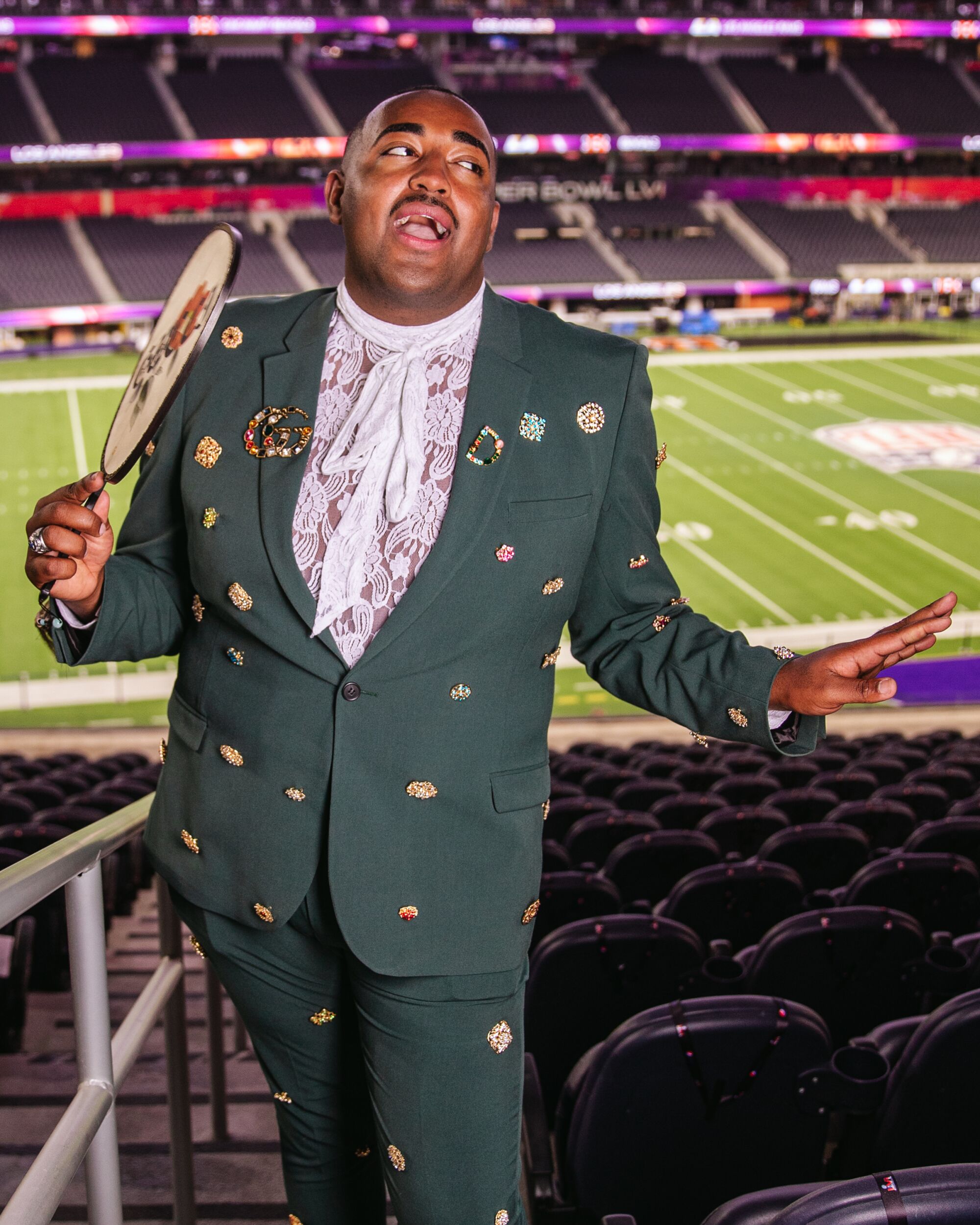 A man in a suit covered with pins and carrying a paper fan poses for a portrait in SoFi Stadium.