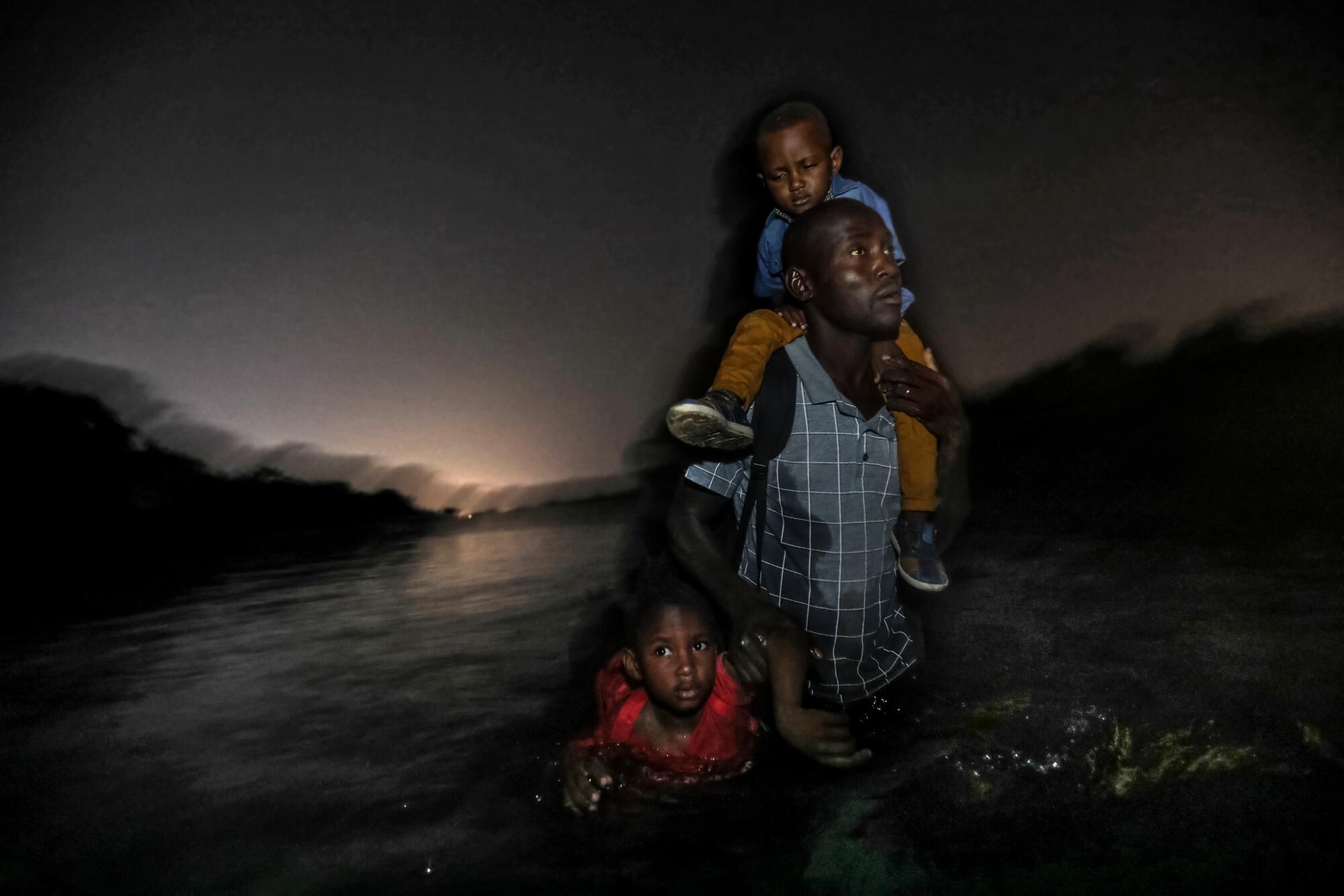 A Haitian man carries one child on his shoulders and holds another by his arm as he wades across the Rio Grande.