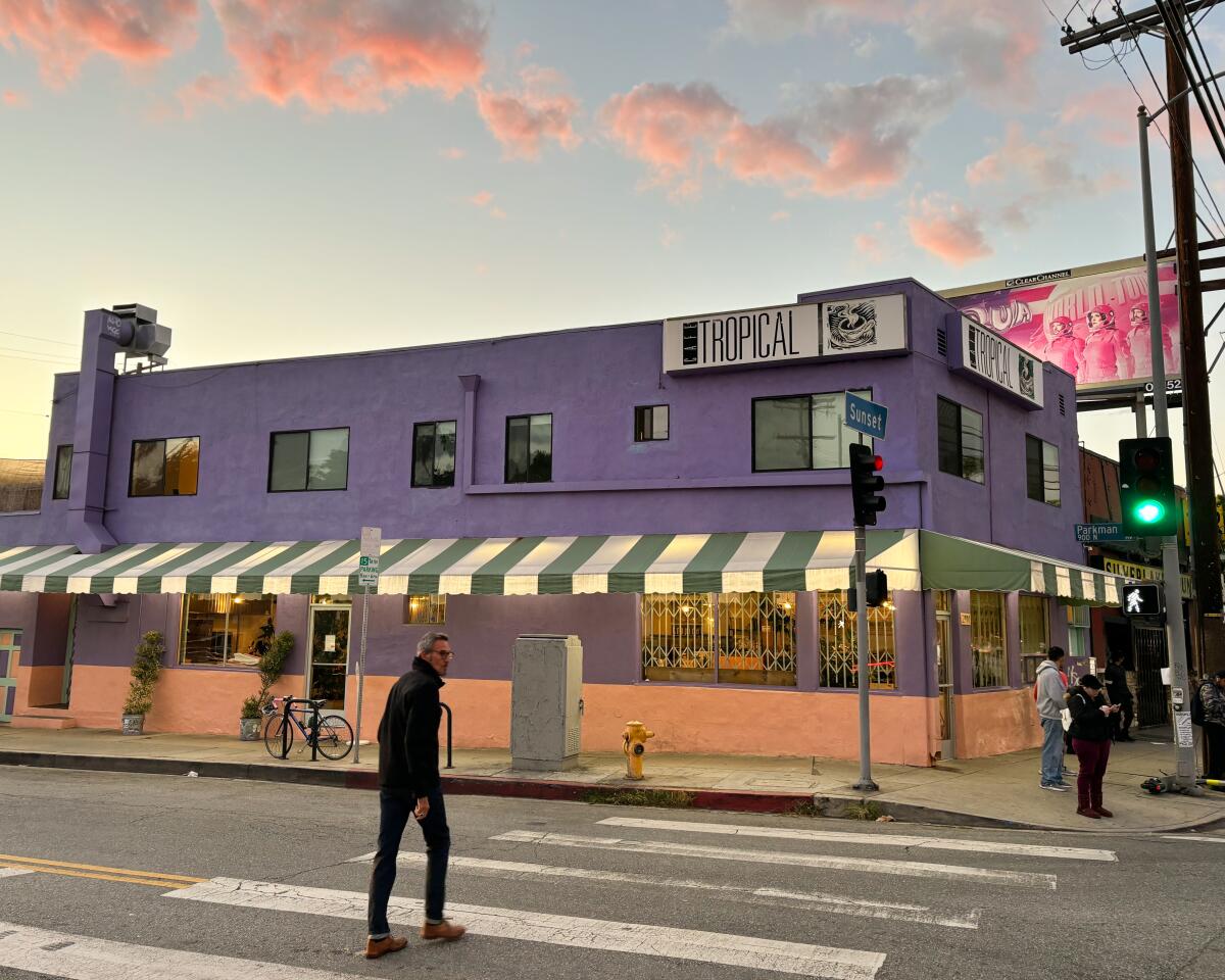 A person crosses the street toward a purple restaurant building under a sky with puffy pink clouds
