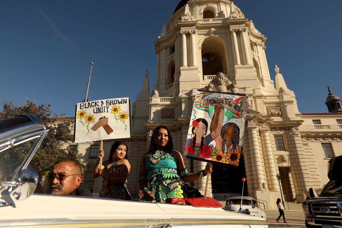 People in a car carry signs that say Black and Brown Unity and Stronger Together in front of Pasadena City Hall