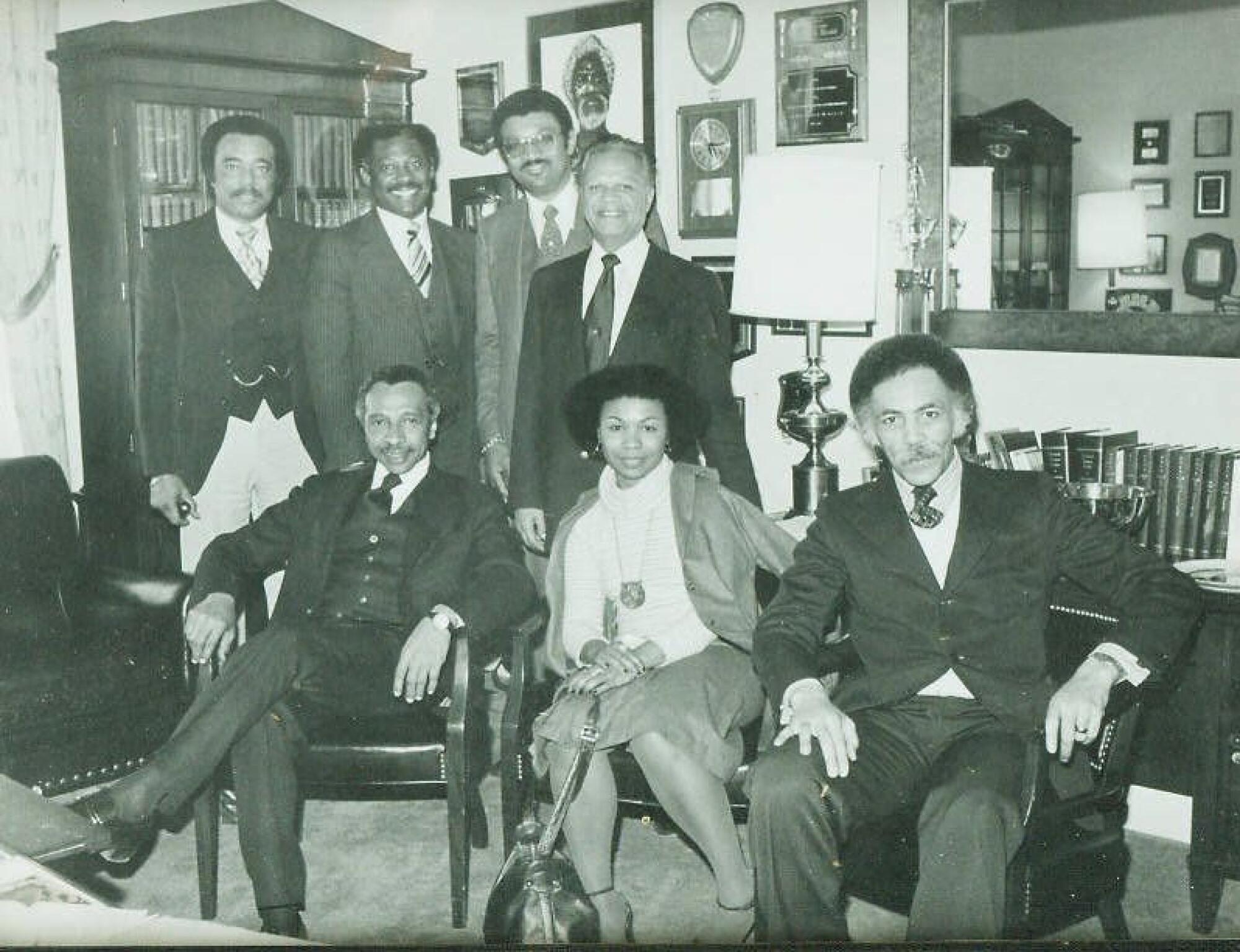 A woman sits in a room with several men who are standing or sitting.