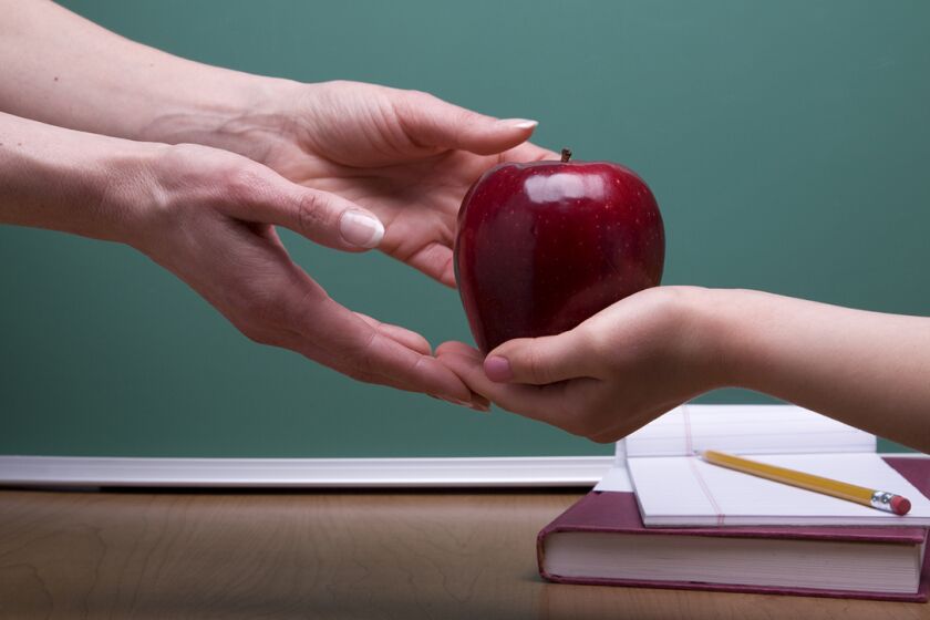 A little girl hands her teacher an apple against a green chalk board.