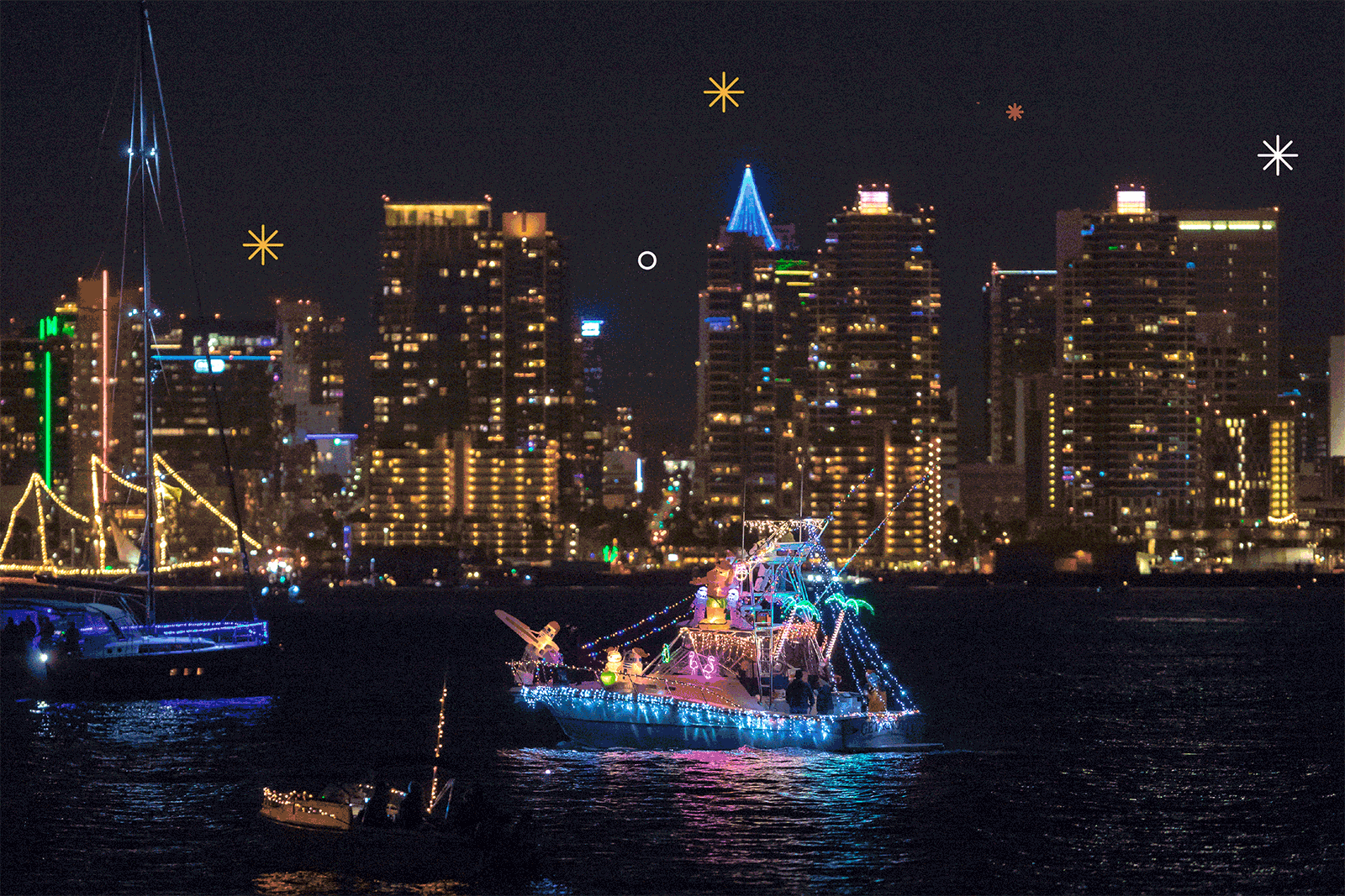 Sailboats, yachts and dinghies decorated with holiday lights sail in water with a cityscape in the background 