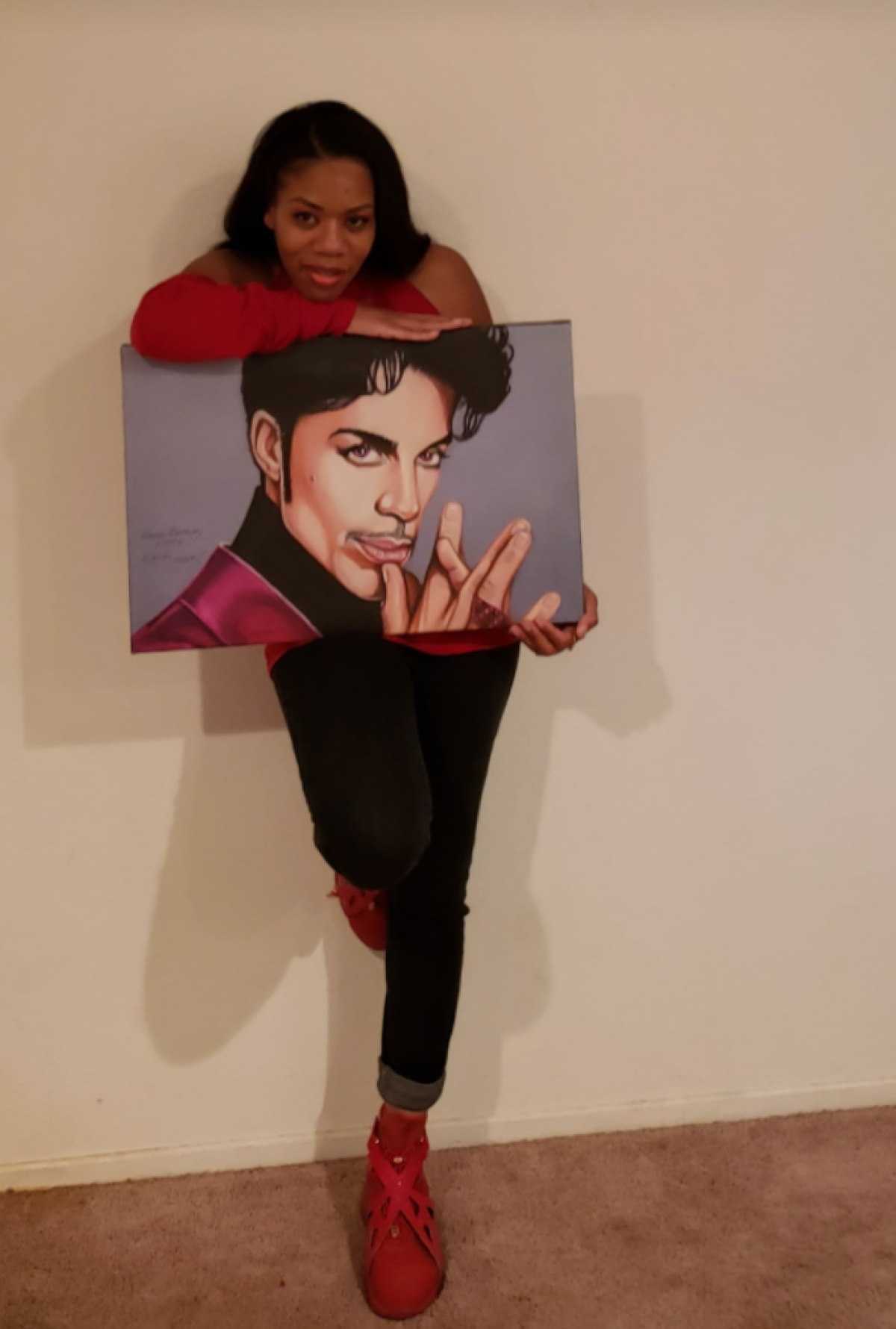 Woman in red shirt, red shoes, and blue jeans holds Prince painting. 