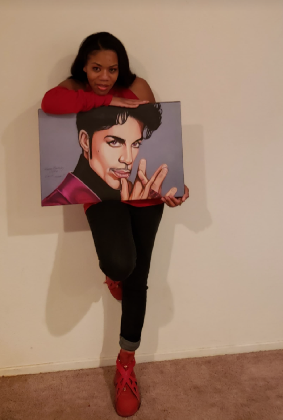 Woman in red shirt, red shoes, and blue jeans holds Prince painting.