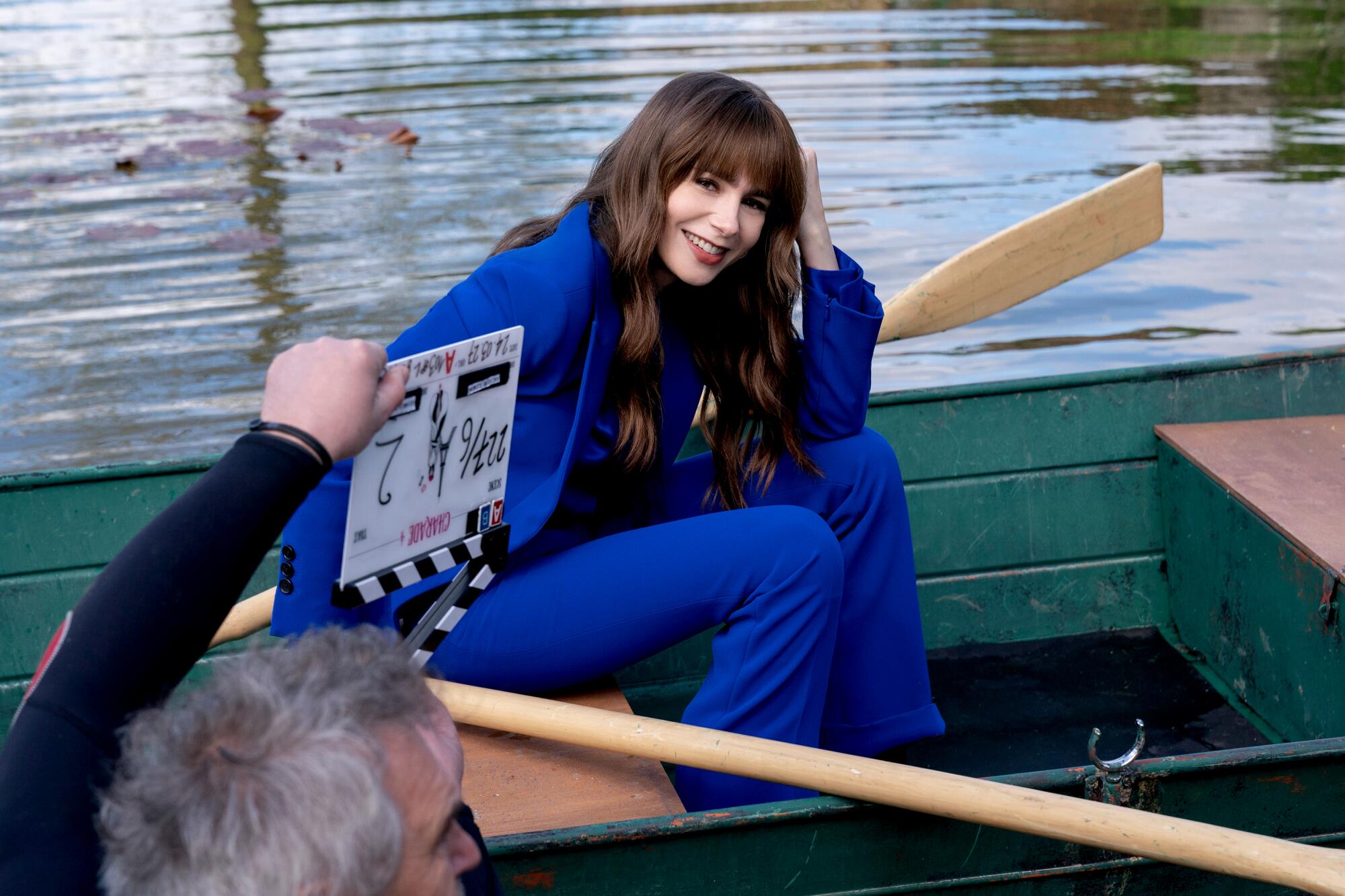 A woman in a blue suit sits near a pair of oars in a rowboat on water.