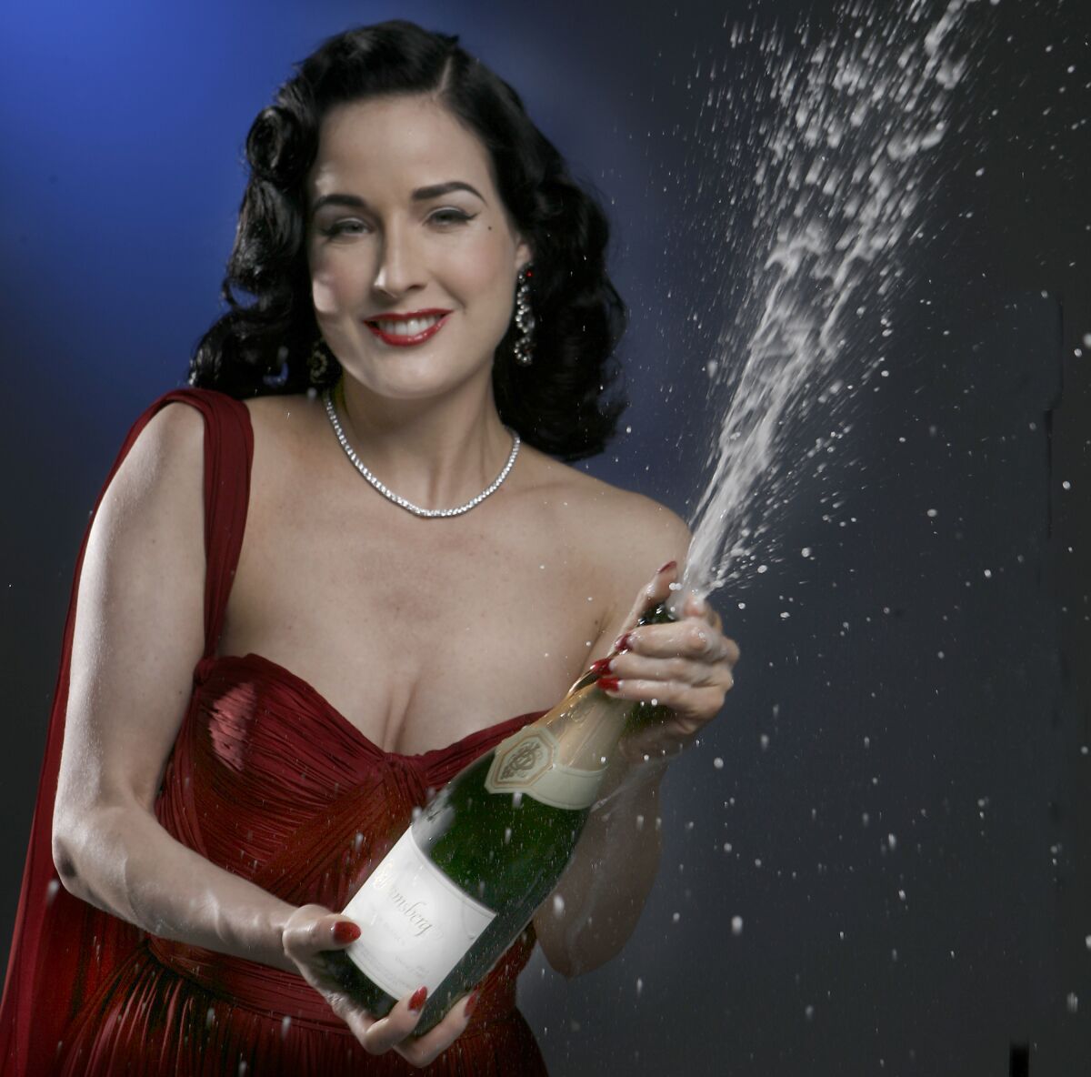 A glamorous woman in a red evening gown pops a bottle of champagne and smiles